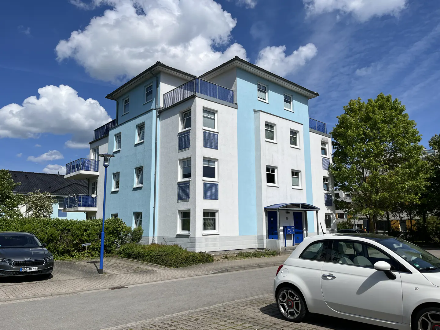 A modern white and light blue apartment building under a blue sky with clouds, with cars parked in the foreground.