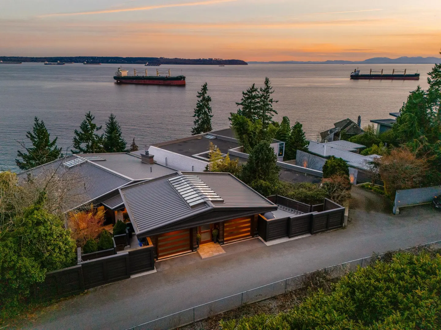 A modern home with a gray roof overlooks the ocean at sunset, with cargo ships visible in the distance.