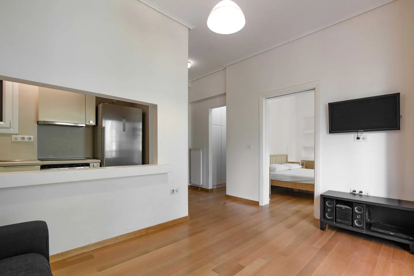 Bright apartment interior with wood floors, white walls, and a view into the kitchen and bedroom. A TV hangs above a black entertainment center.
