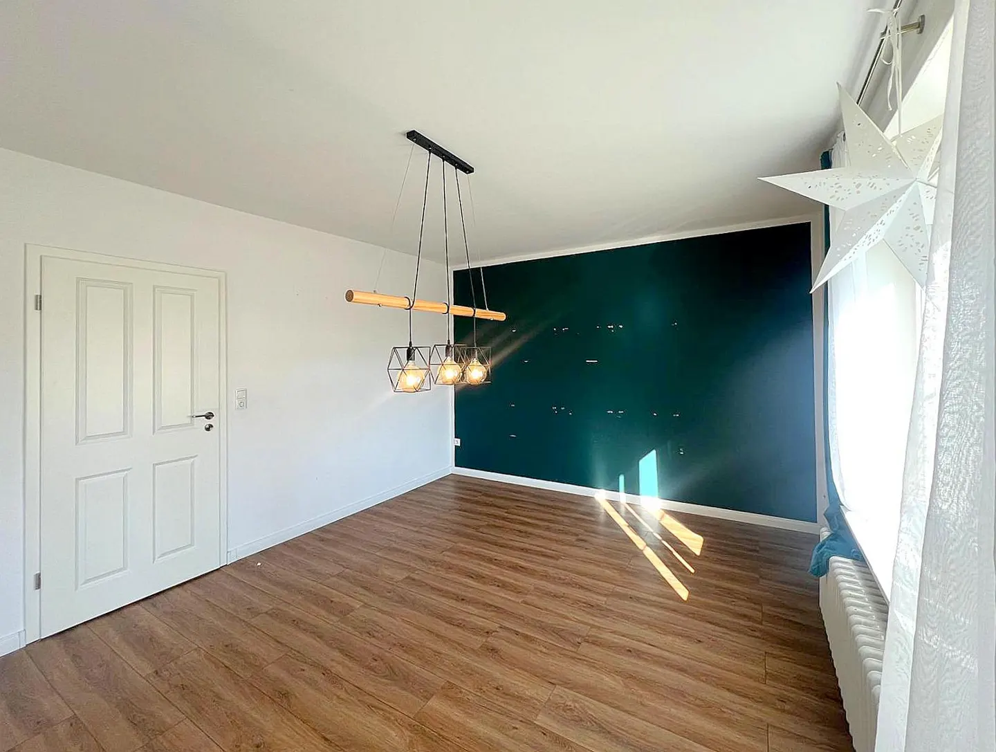 Bright room with wood floors, white walls, and a dark green accent wall. A modern light fixture hangs above, and a white door is on the left.