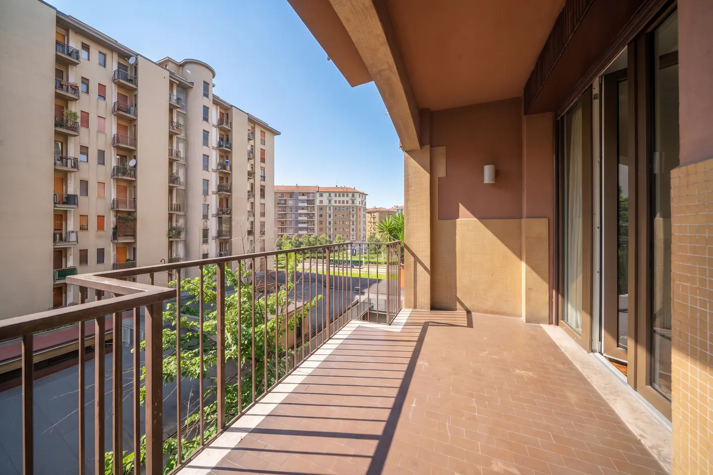 A balcony with brown tile flooring and a metal railing overlooks city buildings and greenery on a sunny day.
