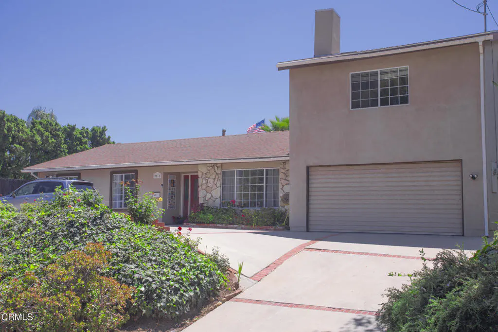 Tan two-story house with a brown roof, a two-car garage, and a driveway lined with red bricks. Bushes and a car are in the front yard.