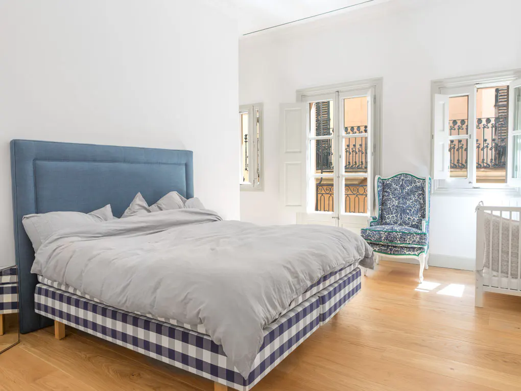Bright bedroom with a blue headboard bed, gray sheets, and a blue and white checkered base. A floral chair and crib sit near open windows.