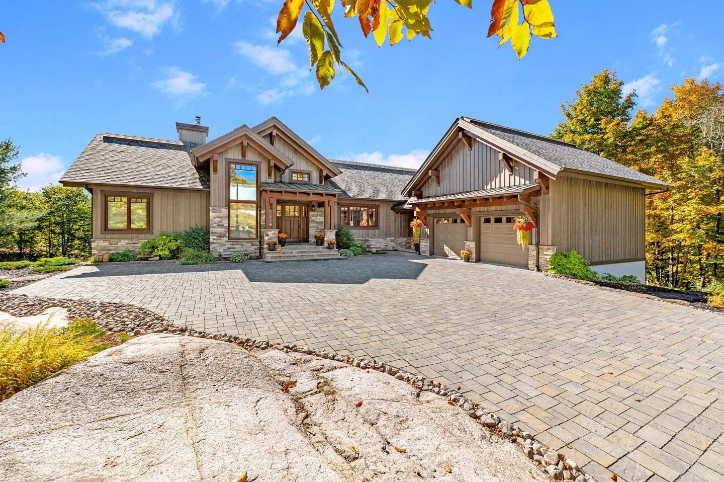 A two-story house with a stone and wood facade, a gray roof, and a paved driveway on a sunny day.