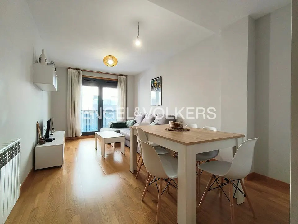 Bright living and dining area with wood floors, white walls, and a balcony. A light gray sofa and a white table with chairs are visible.