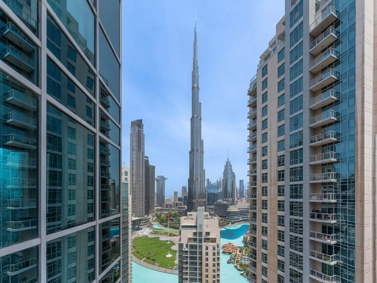 View of the Burj Khalifa in Dubai, framed by two tall buildings. A green park and blue water feature are visible below.