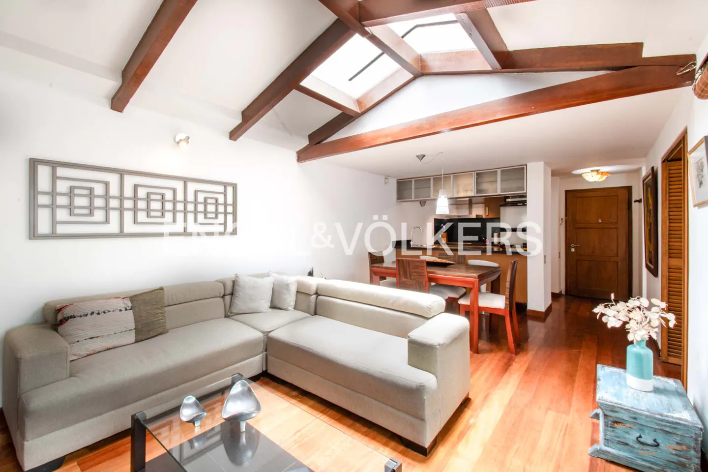 Living room with a beige sectional sofa, glass coffee table, wood floors, and a skylight. A dining table and kitchen are in the background.