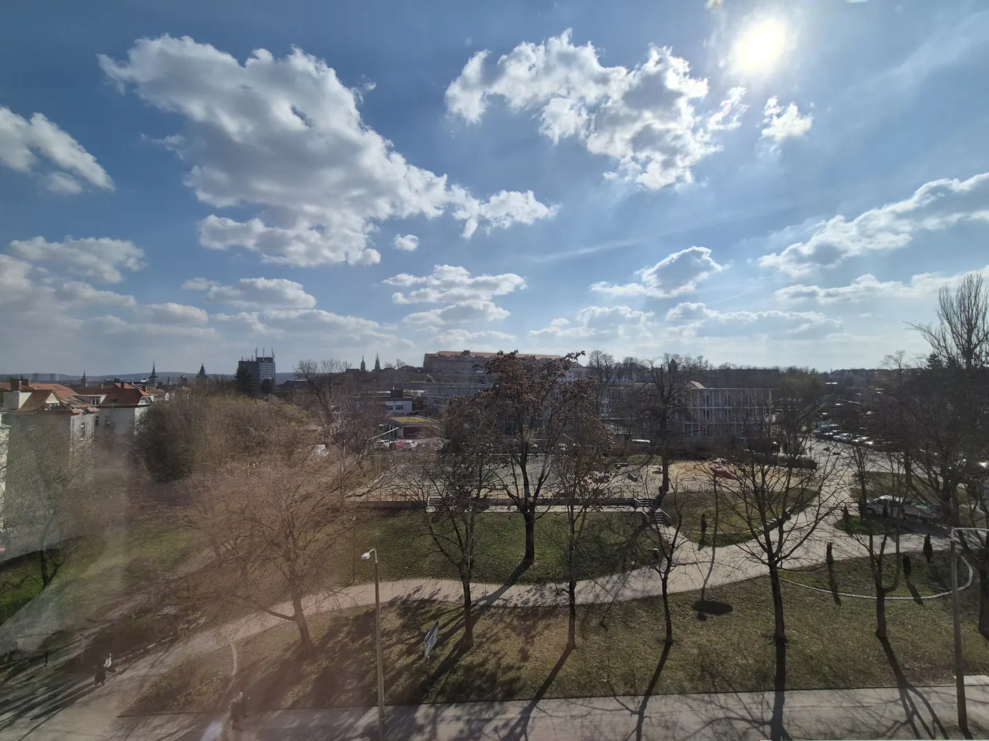 View of a park and city skyline under a blue sky with white clouds and bright sun.