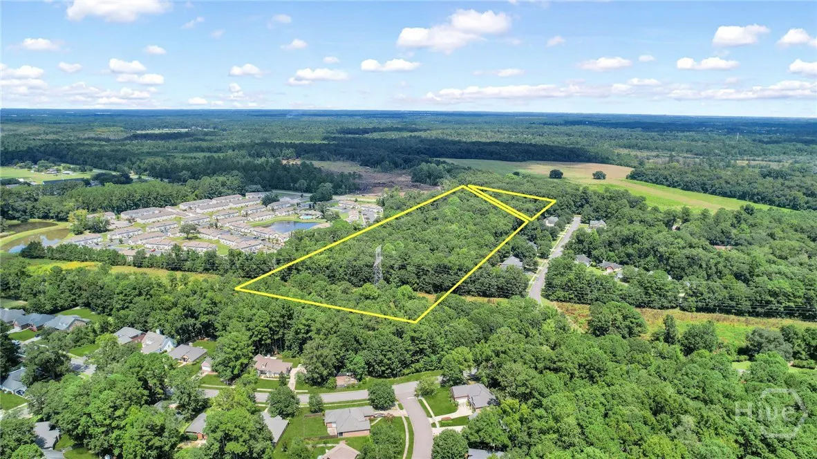 Aerial view of a wooded lot outlined in yellow, surrounded by houses, trees, and a blue sky with white clouds.