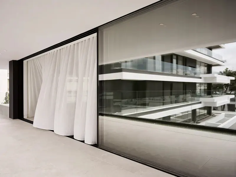 Modern balcony with white sheer curtains and a large sliding glass door reflecting a multi-story building with glass railings.