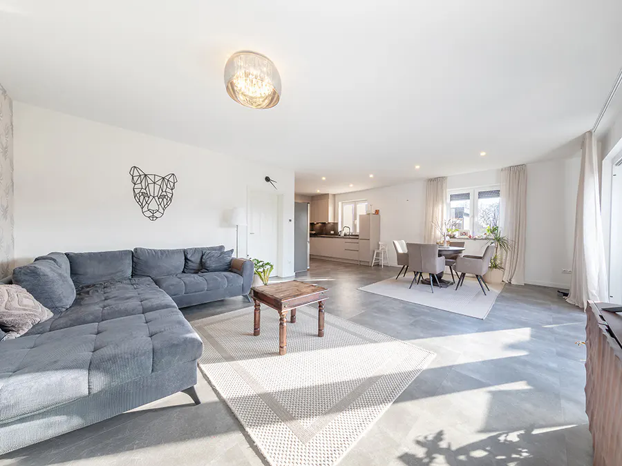 Modern open-plan living room with a grey sectional sofa, wooden coffee table, and a dining area with a kitchen beyond.
