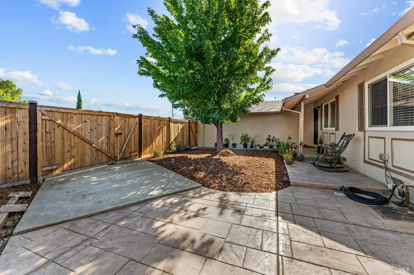 Backyard patio with stamped concrete, a wood fence, a large tree, mulch area, and a rocking chair on the porch.