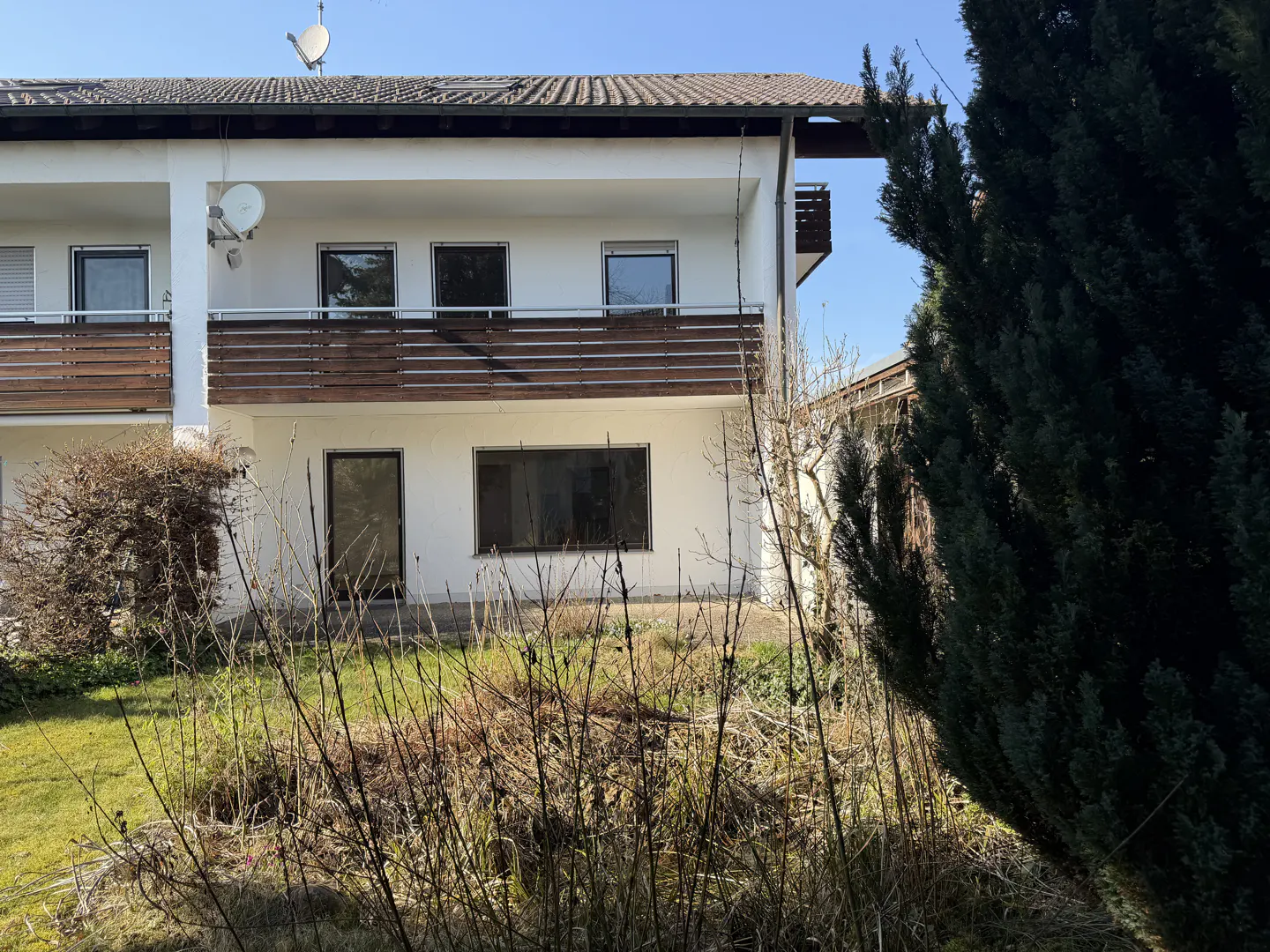 Two-story white building with brown balconies, visible windows, and a satellite dish on the roof. Overgrown yard in front.