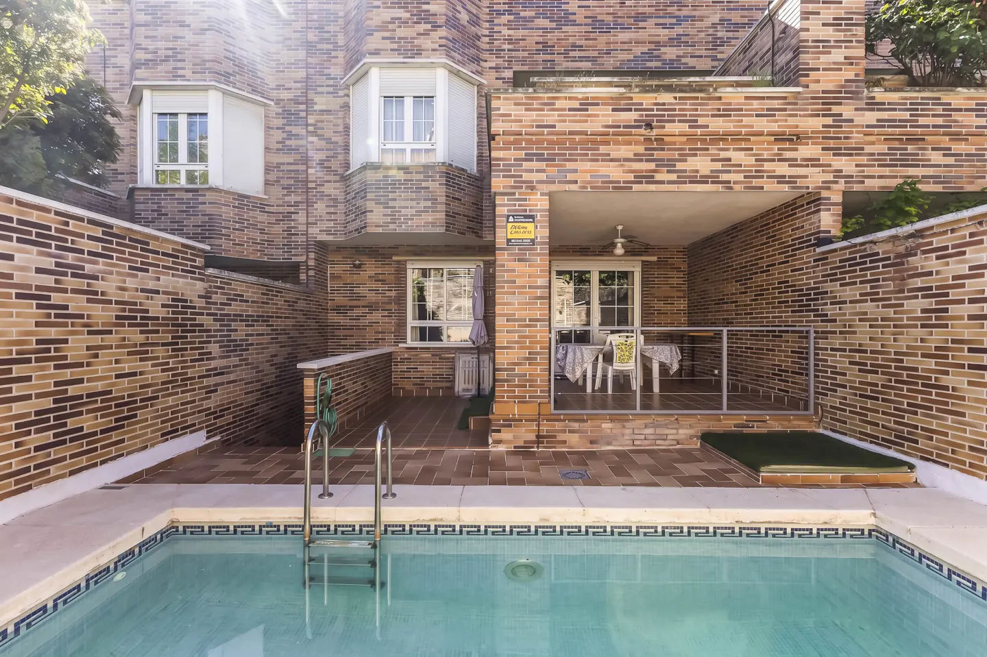 Exterior view of a brick house with a pool. The pool has a metal ladder and blue tile trim. A covered patio has a table and chairs.
