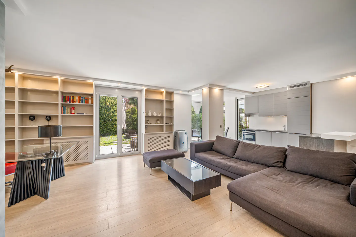 Bright, open-concept living space with a brown sectional sofa, glass coffee table, and light wood floors. Bookshelves and a modern kitchen are visible.