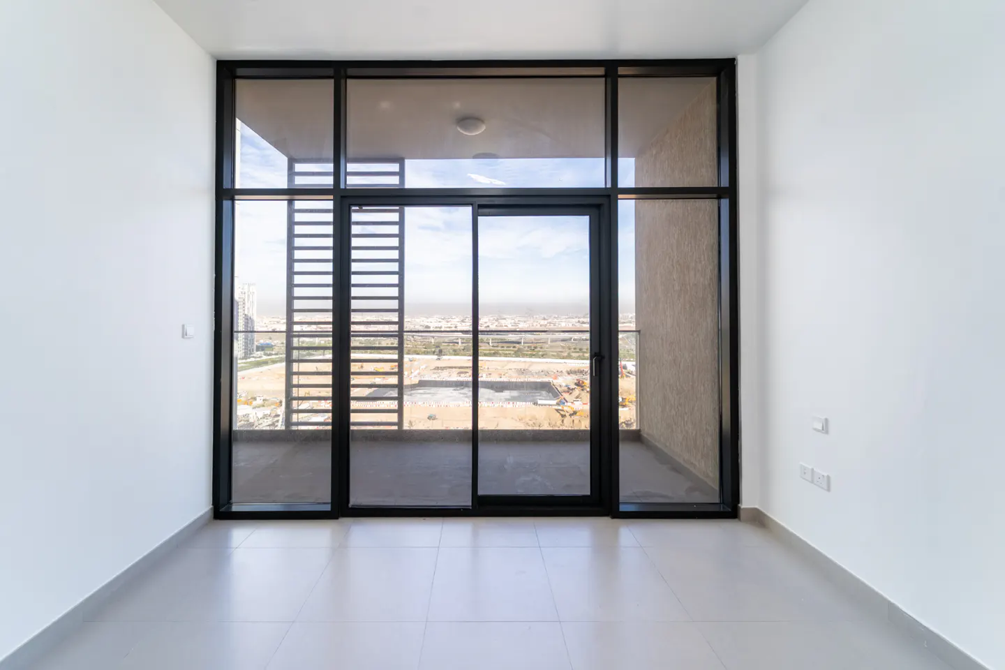 Empty room with white walls and tile floor. Black framed sliding glass doors lead to a balcony with city views.