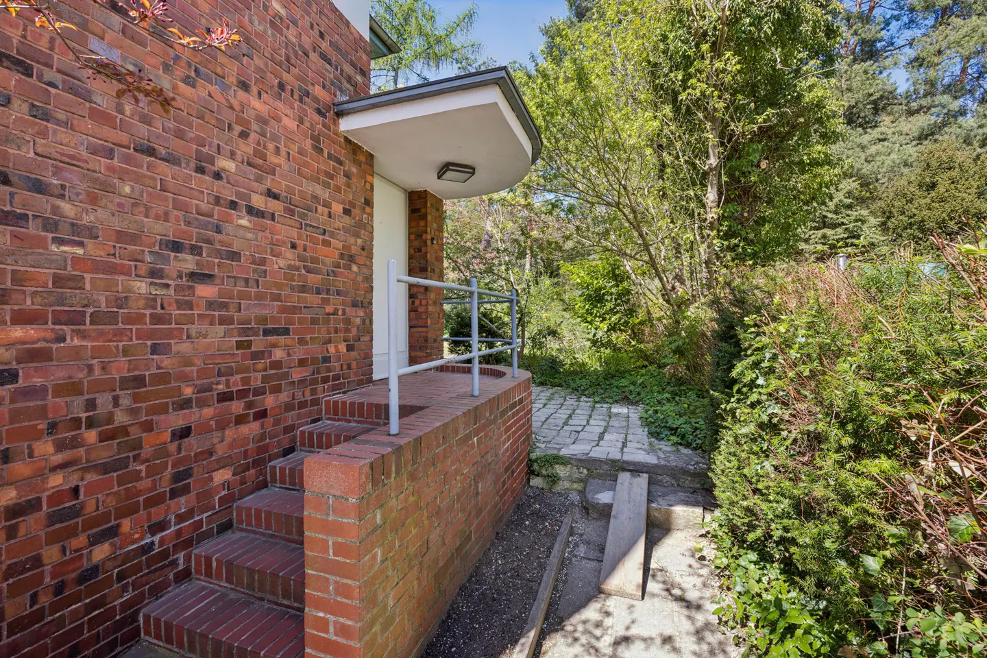 Brick house entrance with steps, a white door, and a gray handrail. Lush greenery surrounds a stone path leading away from the house.
