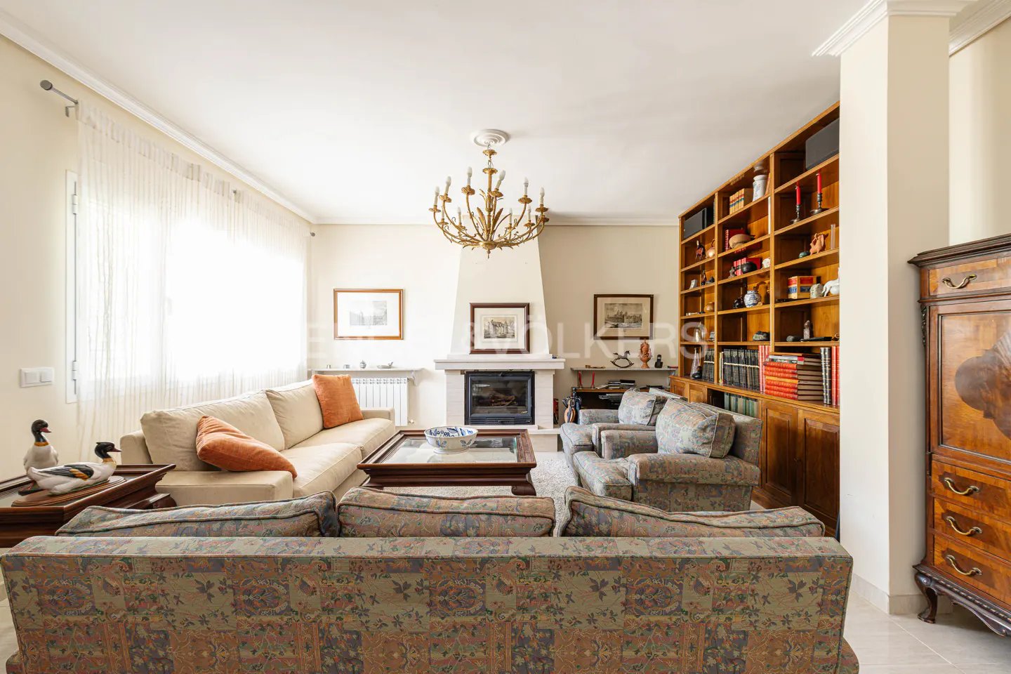 Bright living room with a beige sofa, patterned armchairs, a fireplace, and a wooden bookcase. A chandelier hangs from the ceiling.