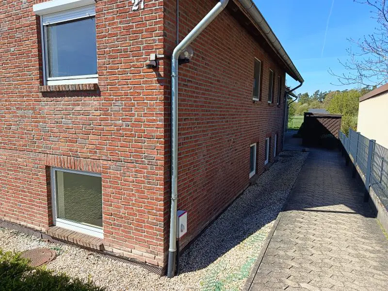 Exterior view of a red brick house with white-framed windows and a silver gutter. A paved driveway runs alongside the house.