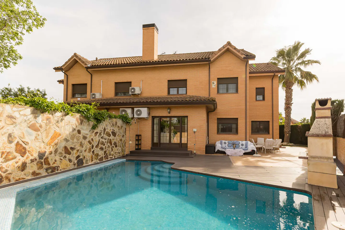 Backyard view of a two-story brick house with a pool, stone wall, and patio furniture. A palm tree stands tall in the background.