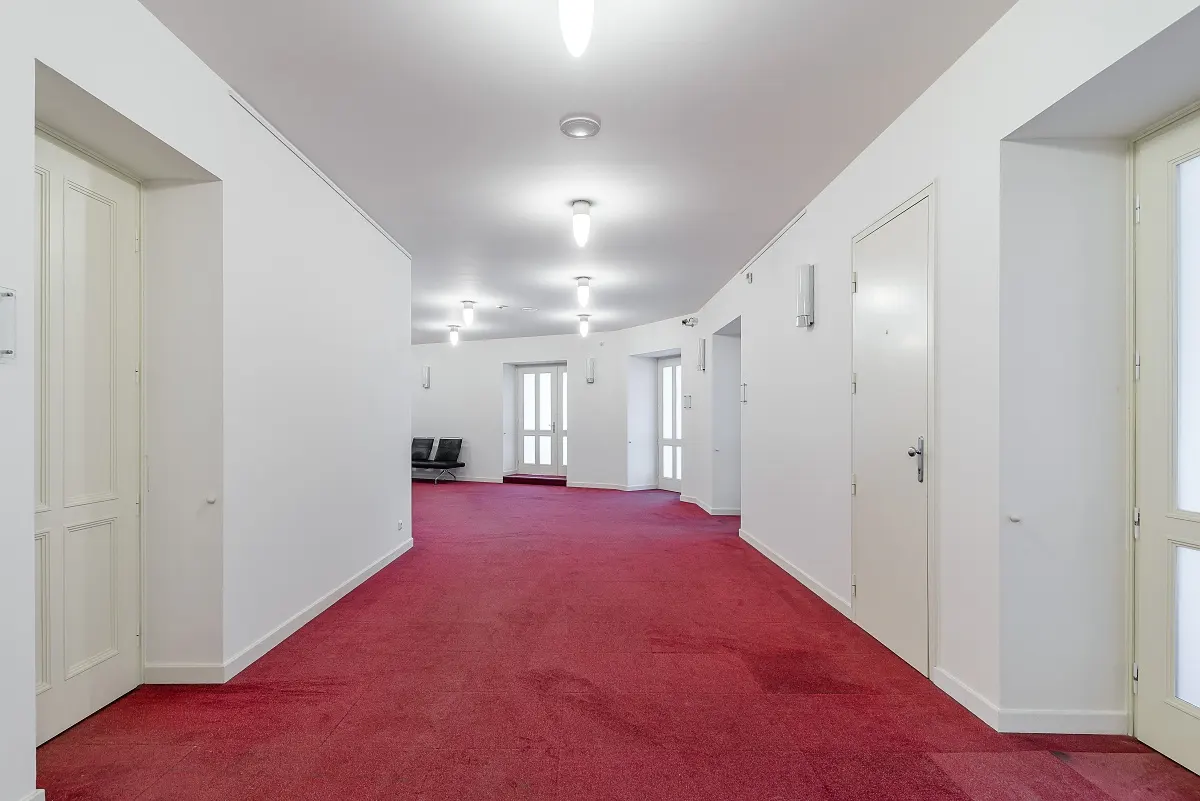 Hallway with red carpet and white walls. Doors line the walls, and lights are on the ceiling. Two black chairs are in the distance.