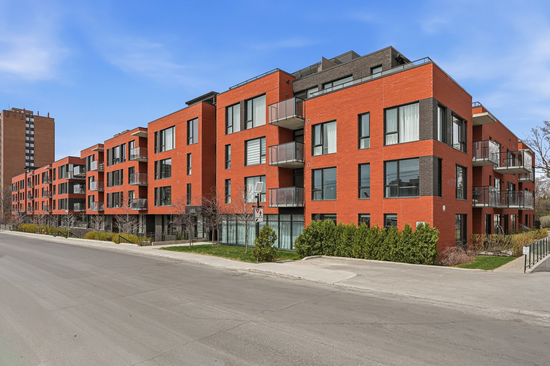 A modern, multi-story red brick apartment building with black framed windows and balconies under a blue sky.