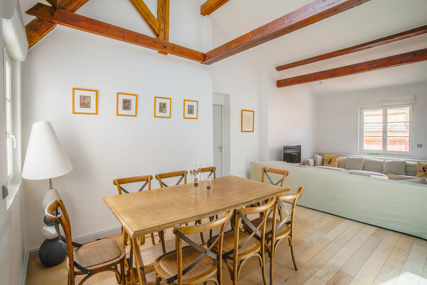 Bright, airy dining room with wood table, chairs, and floors. White walls, exposed beams, and framed art. Sofa in background.