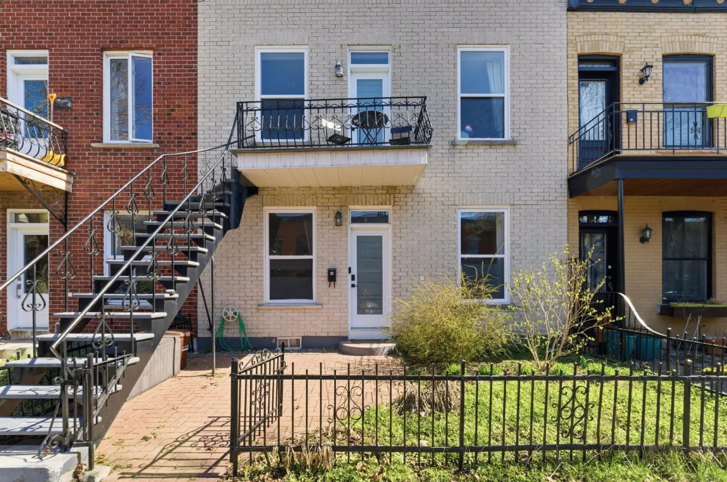 Exterior view of three brick row houses with black iron stairs and balconies. A black fence surrounds a small yard.