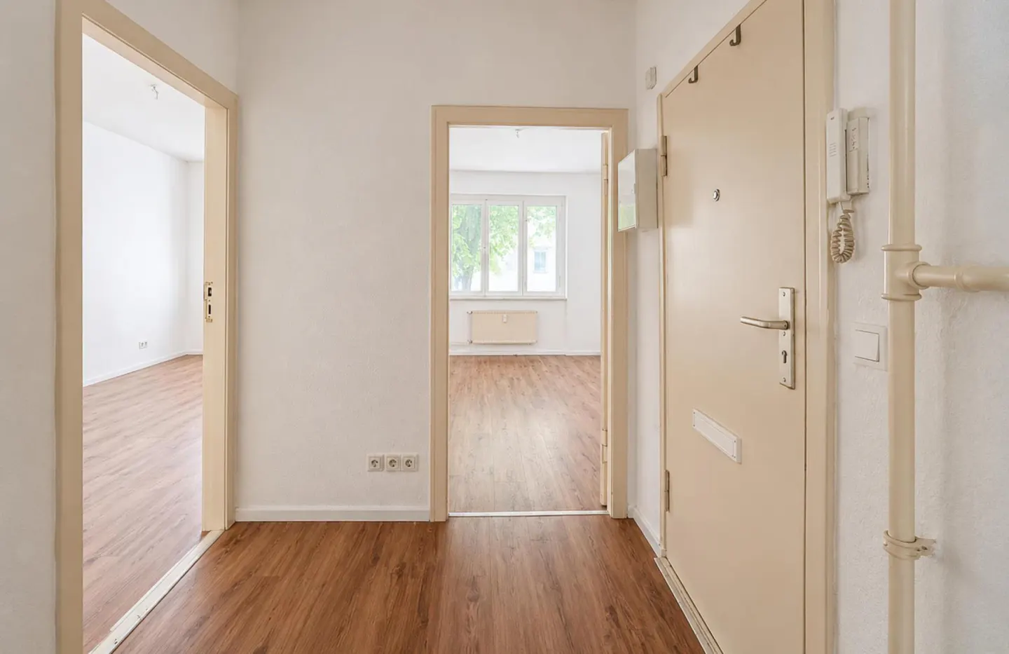 A bright hallway with wood-look flooring, a beige entry door, and two open doorways leading to empty rooms with windows.