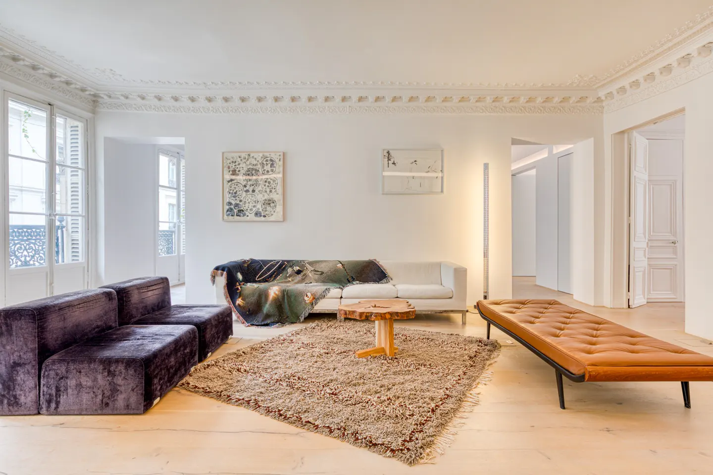 Bright living room with white walls, ornate ceiling, purple sofa, white couch with blanket, and brown leather bench.