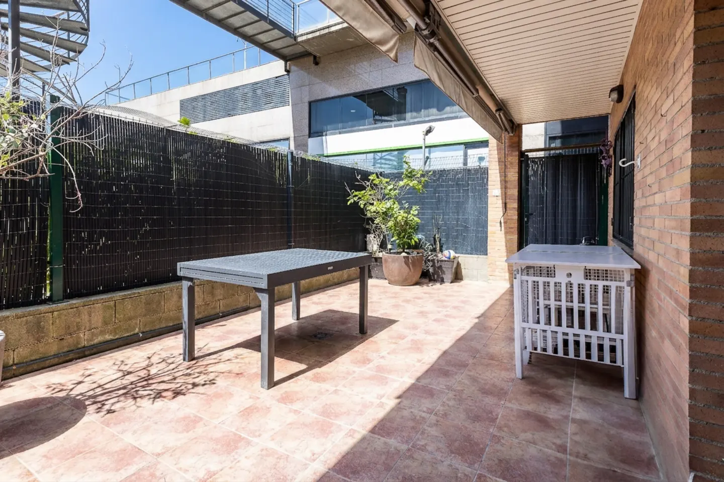 Outdoor patio with a gray table, white table, potted plants, and a black fence. The floor is tiled in a terracotta color.
