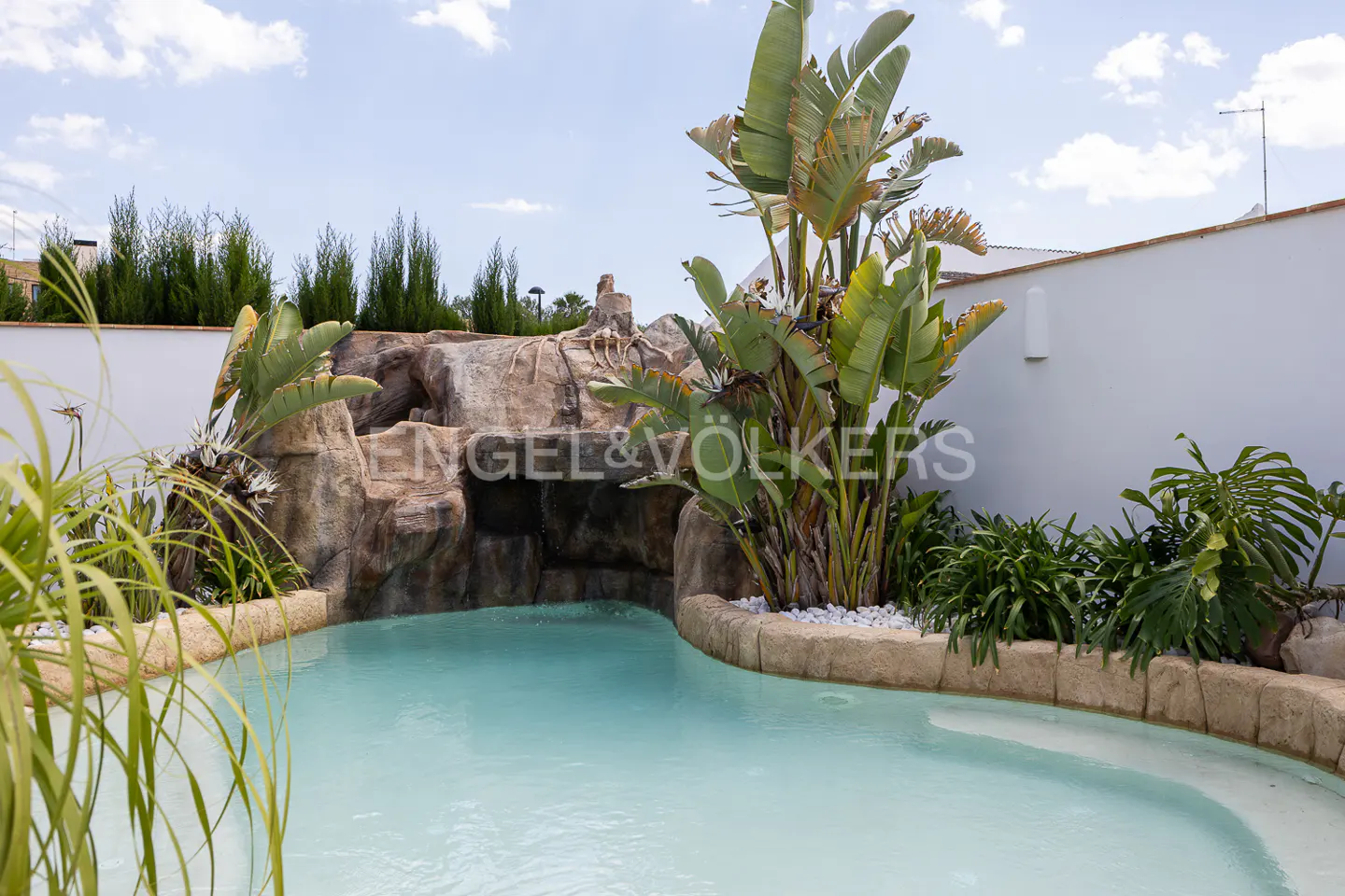 A backyard pool with a rock waterfall feature and tropical plants. The water is light blue and the sky is partly cloudy.