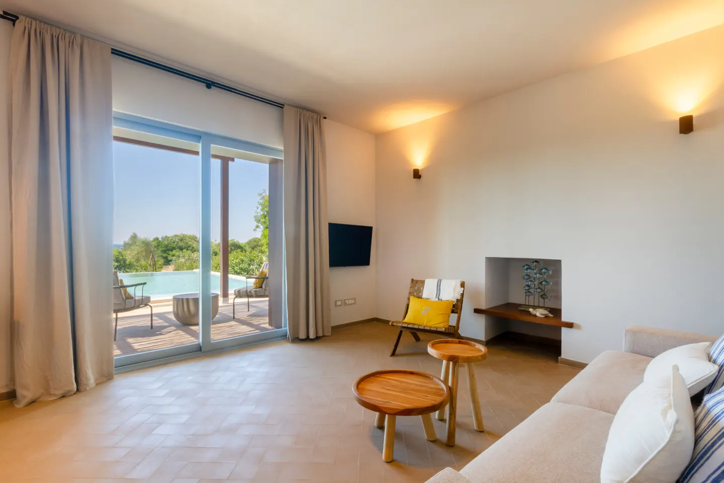 Modern living room with beige sofa, wooden coffee tables, and sliding glass doors opening to a sunny pool patio.