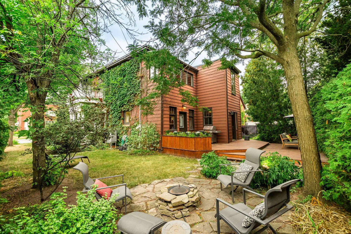 Backyard view of a brown house with a stone patio, fire pit, and gray chairs surrounded by lush green trees and grass.