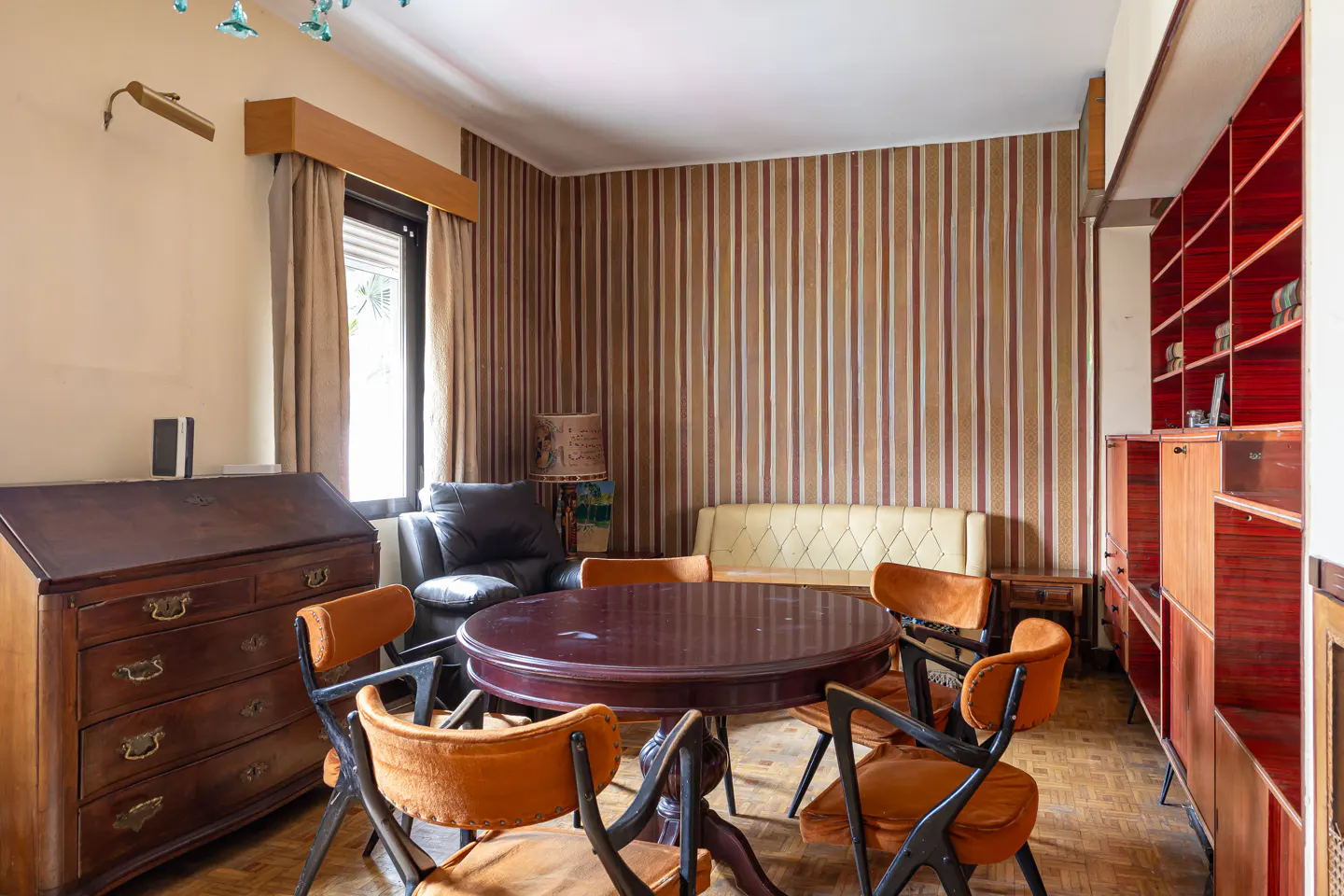 A vintage-style room with a round table, orange chairs, and a wooden desk. Striped wallpaper and a red bookshelf add to the retro aesthetic.