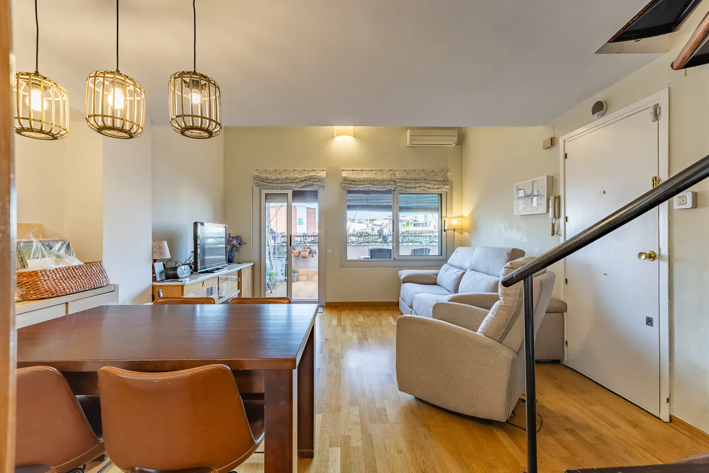 Bright living room with wood floors, a dining table, a beige sofa, and a door leading to a balcony. Three pendant lights hang above the table.