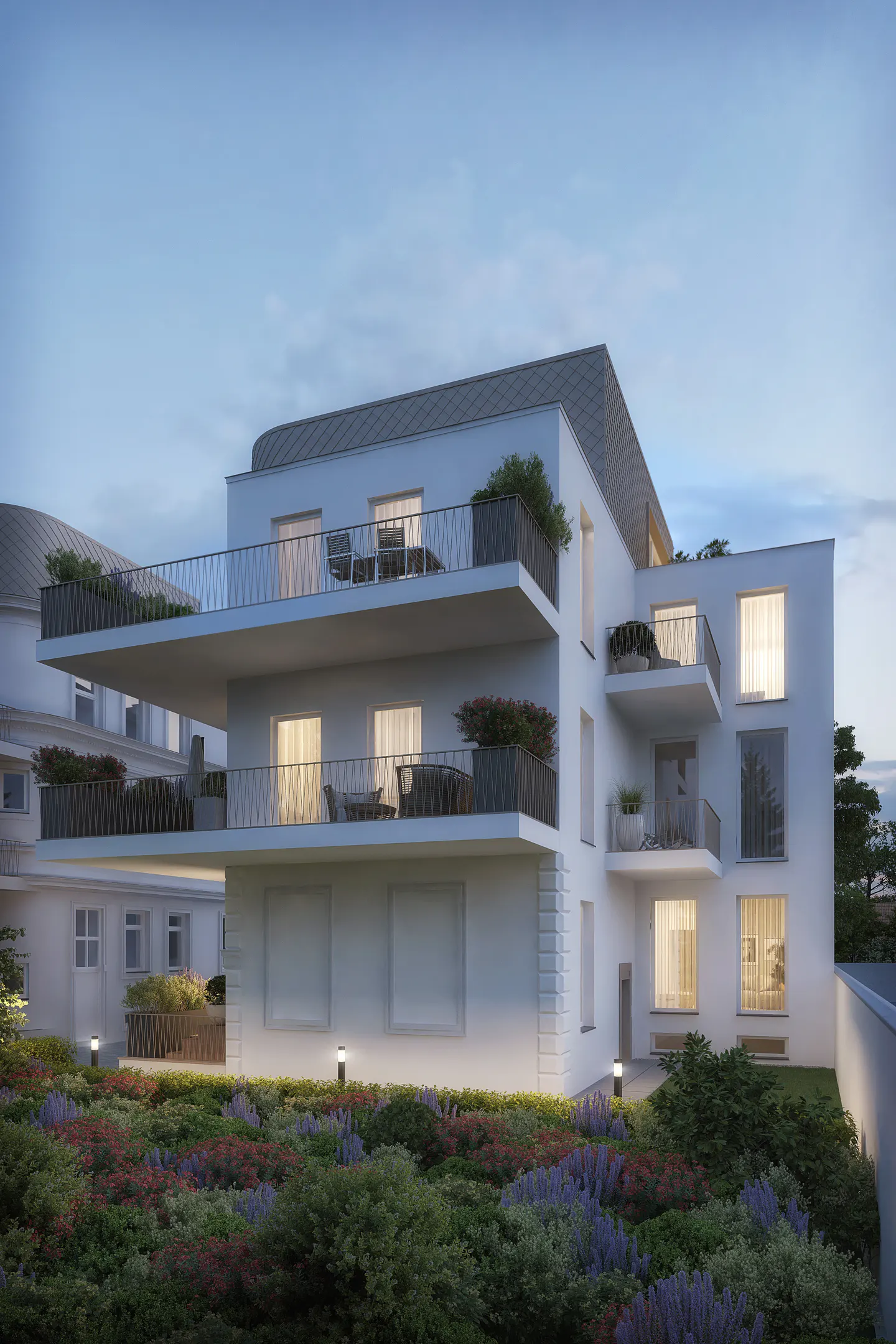 Modern white apartment building at dusk with illuminated balconies, large windows, and a lush garden in the foreground.