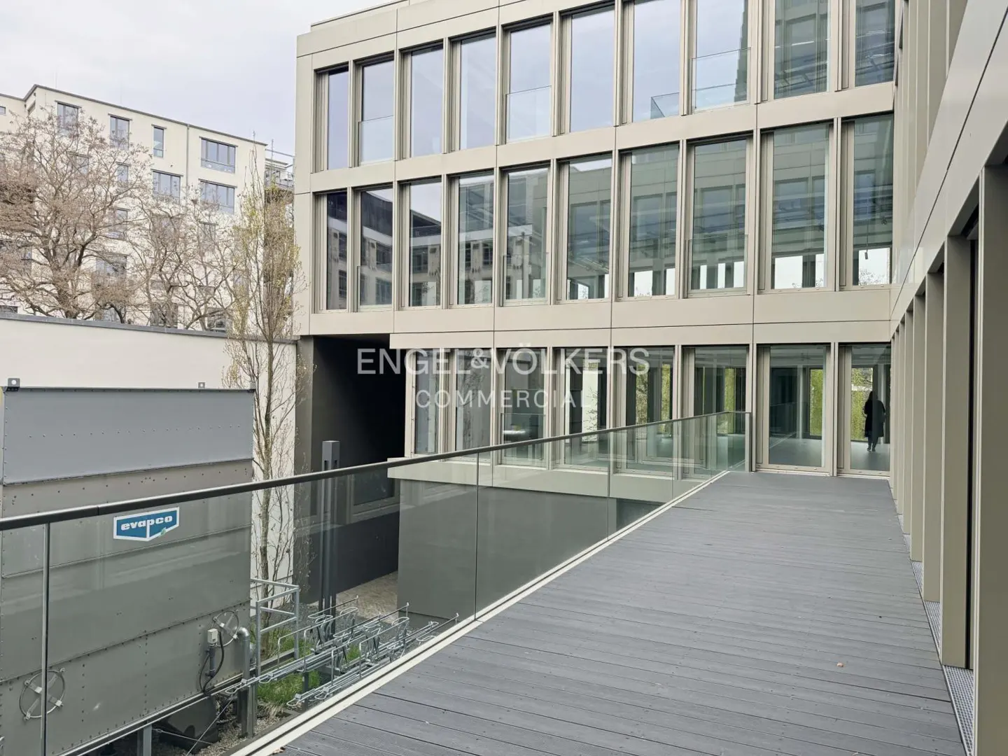 Exterior view of a modern commercial building with large windows and a gray deck with glass railings. The Engel & Volkers logo is visible.