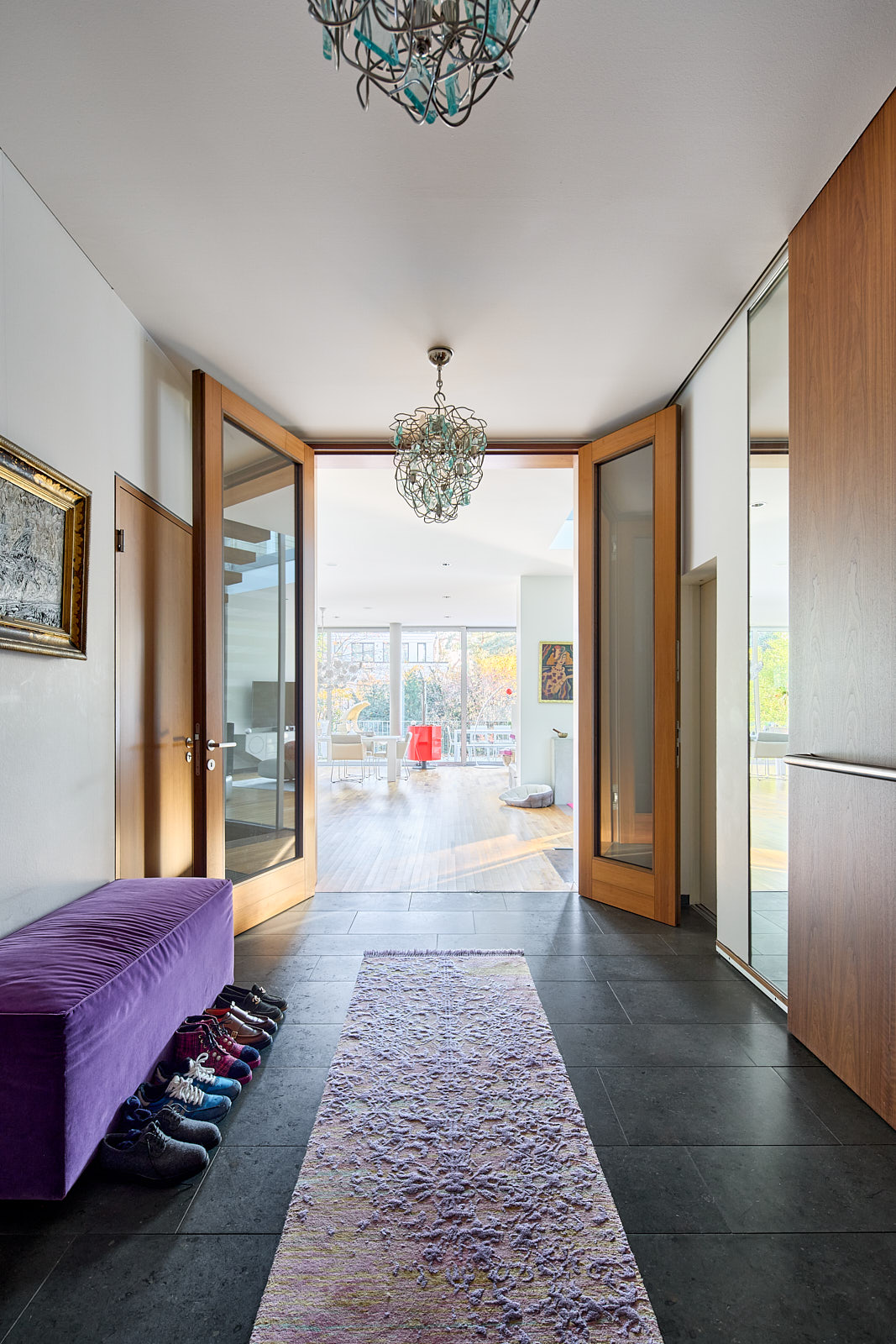 A modern home's foyer with a purple bench, shoes, and a textured rug on dark tile. Light streams in through glass doors.