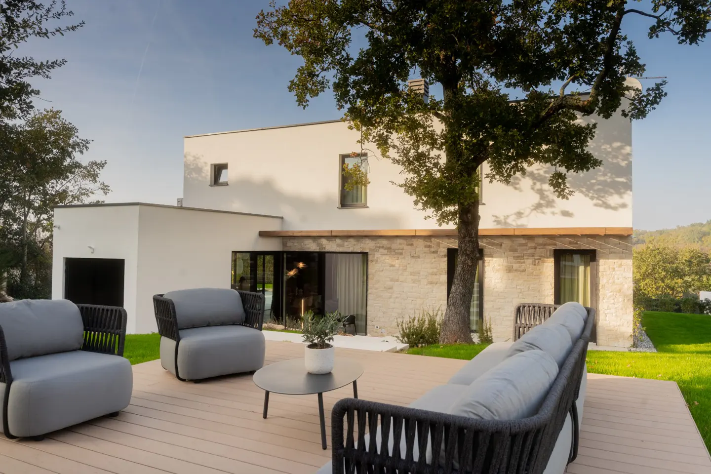 Modern home exterior with a wood deck, gray outdoor furniture, and a large tree. The house has white and stone walls.
