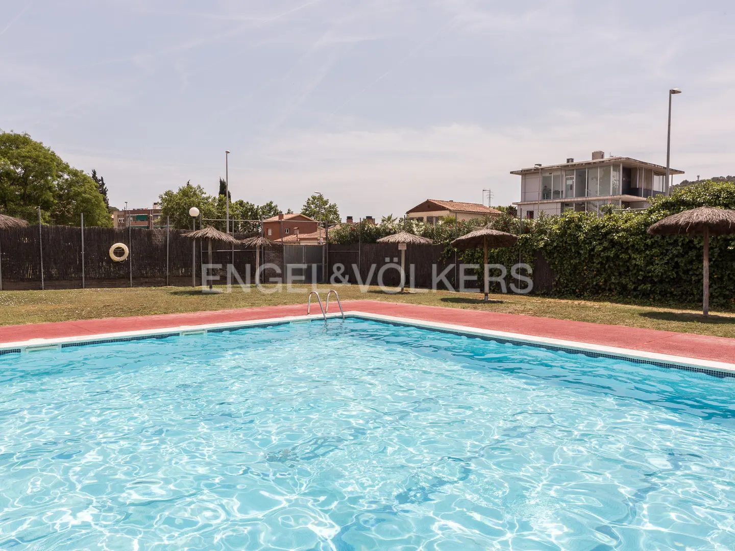Outdoor swimming pool with blue water, red border, and straw umbrellas in a grassy area with a fence and houses behind.
