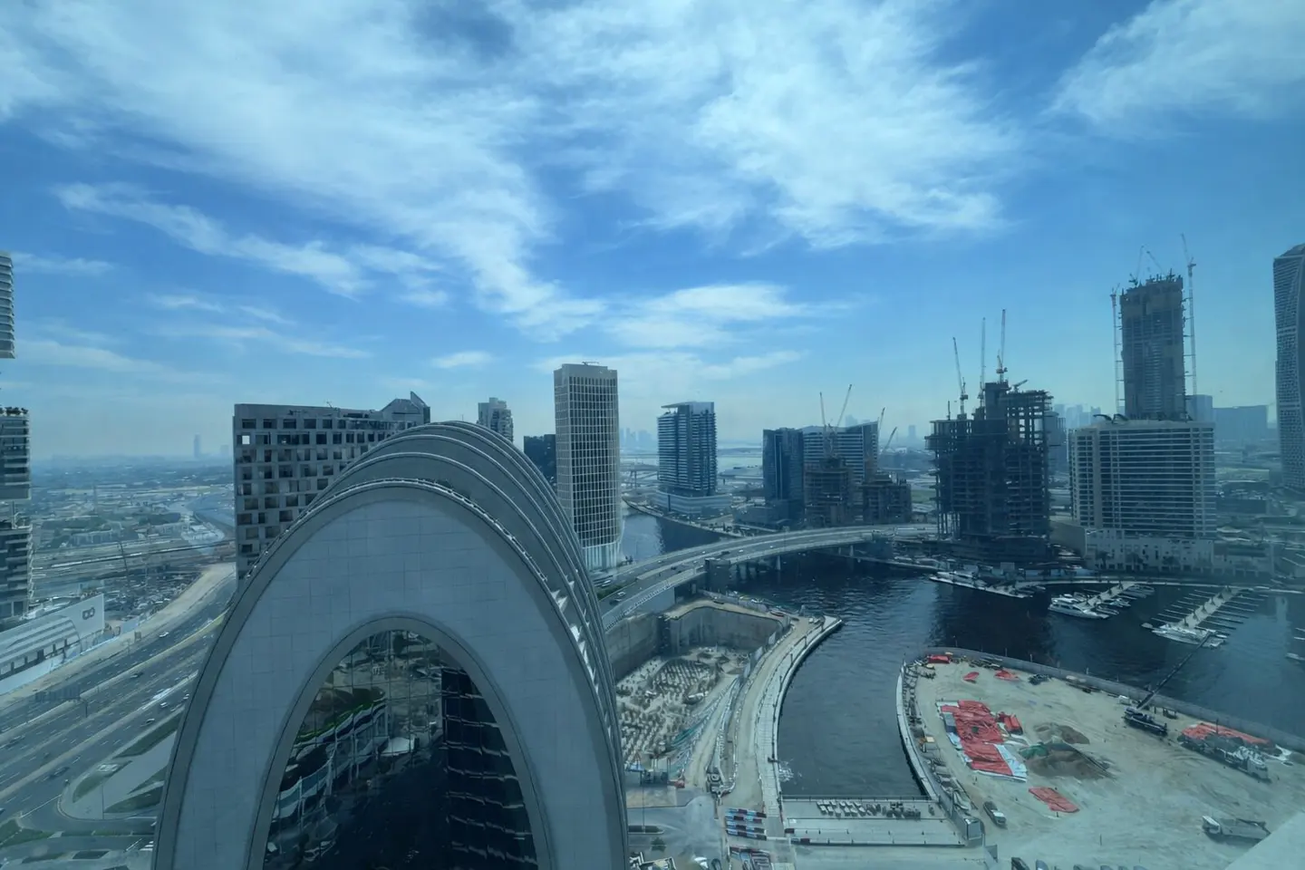 Cityscape view from a high-rise, featuring modern buildings, a river, and a bridge under a blue sky with scattered clouds.