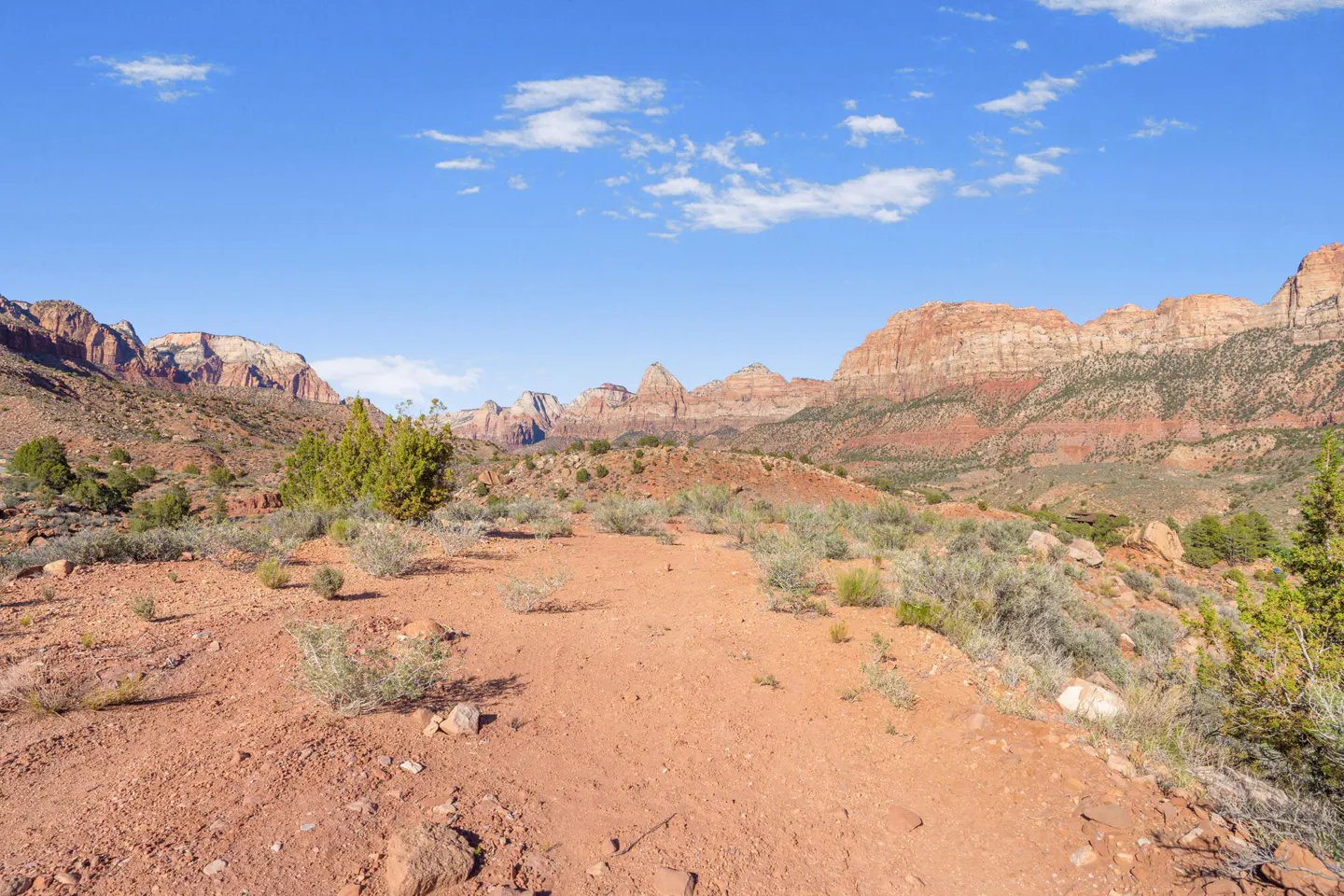 Desert landscape with a red dirt path leading to layered mountains under a blue sky with scattered clouds.
