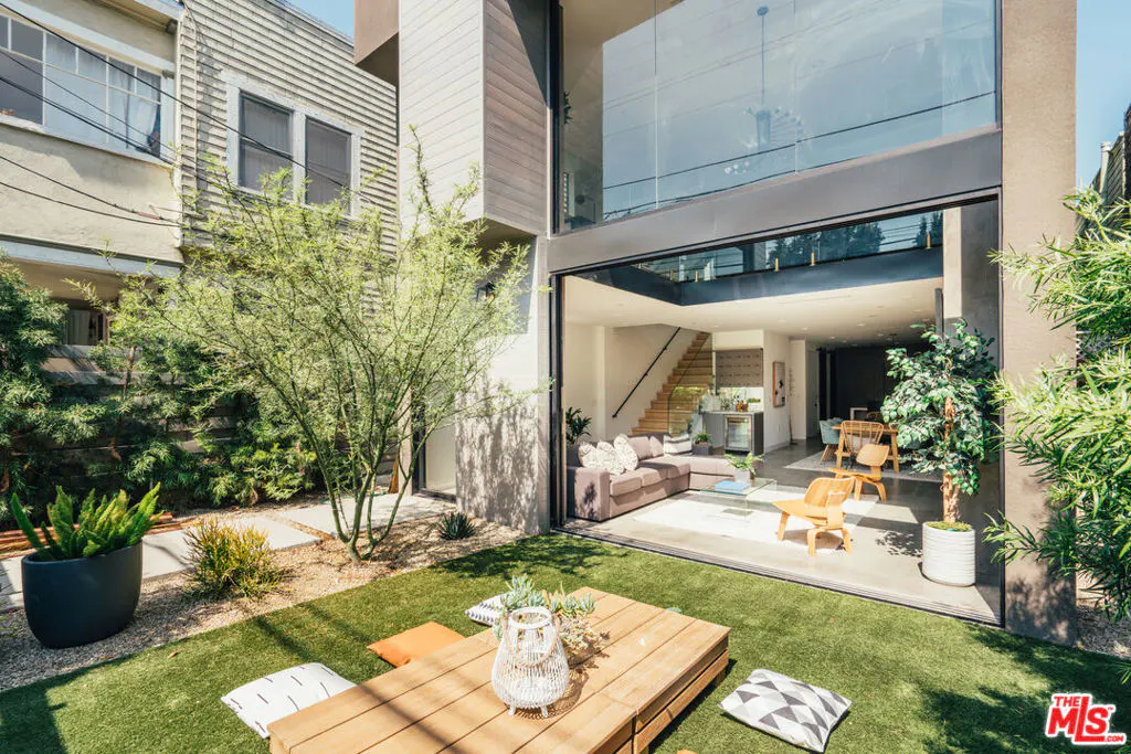 Outdoor patio with a wooden table, pillows, and a view into a modern home with a large glass wall.