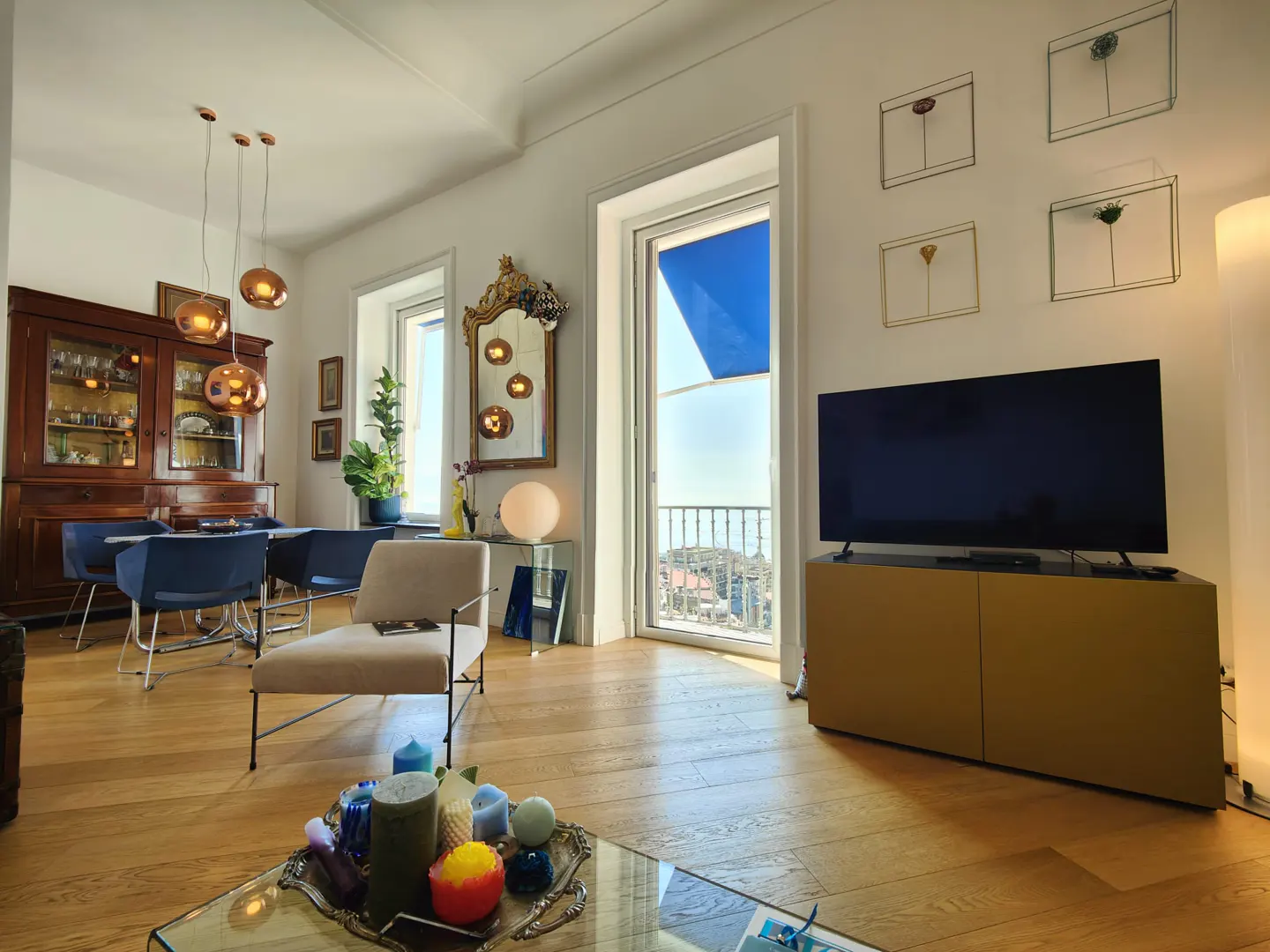 Bright living room with wood floors, a dining table, and a TV. A balcony with a blue awning is visible through a window.