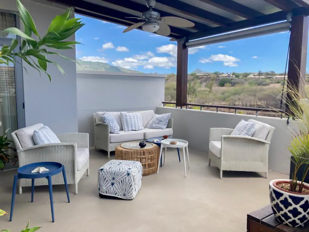Outdoor patio with white wicker furniture, blue accents, and a view of rolling hills under a blue sky. A ceiling fan hangs above.