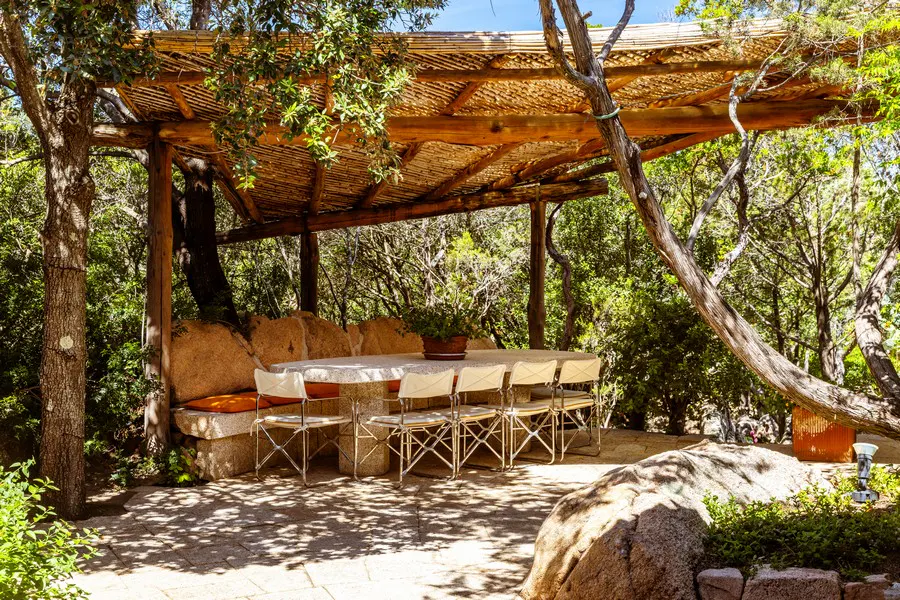 Outdoor dining area with a stone table, white chairs, and a thatched roof pergola surrounded by trees.