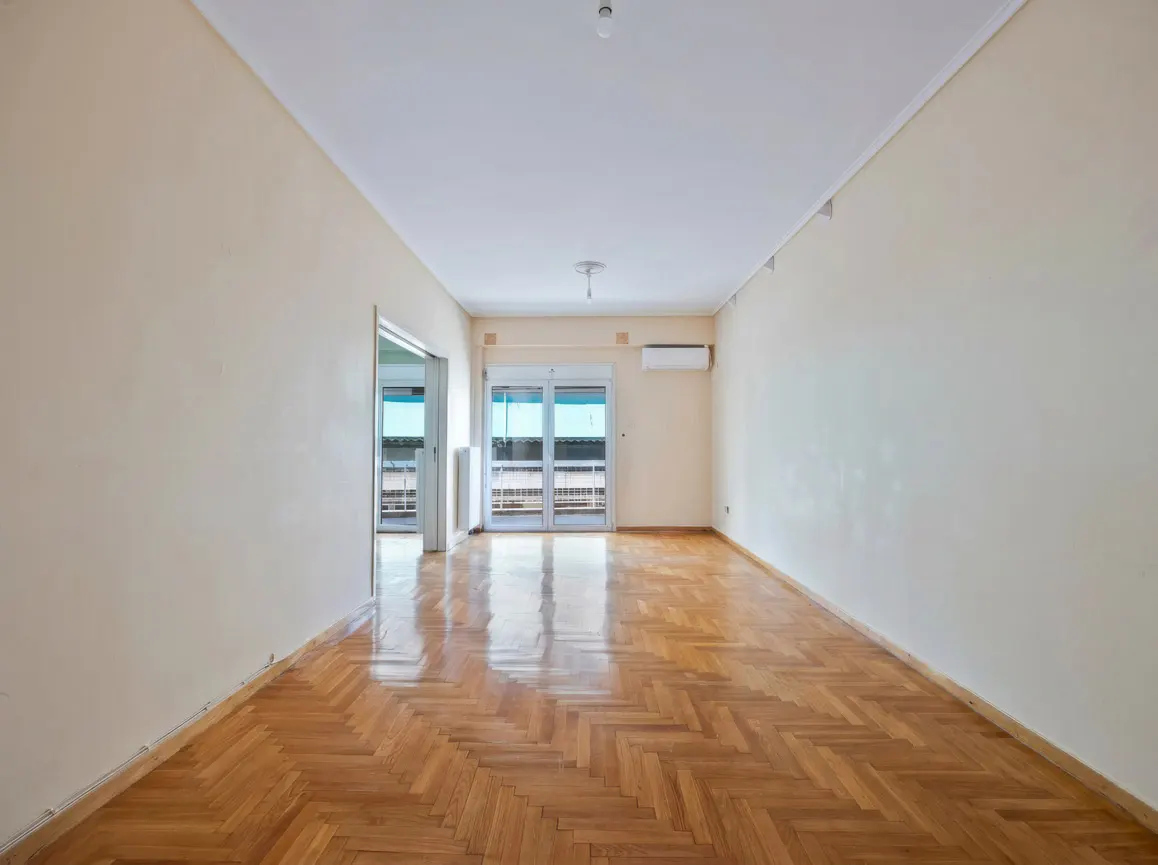 Empty room with light-colored walls, polished herringbone wood floors, an air conditioner, and glass doors to a balcony.