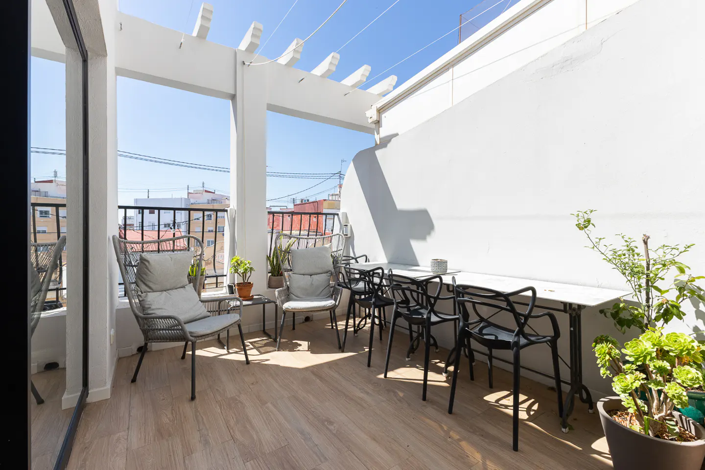 A bright balcony with a white pergola, wood-look flooring, and a white wall. There are gray chairs, a white table with black chairs, and potted plants.