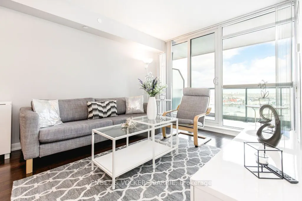 Bright living room with a gray sofa, patterned rug, and a glass coffee table. A chair faces a large window with a city view.